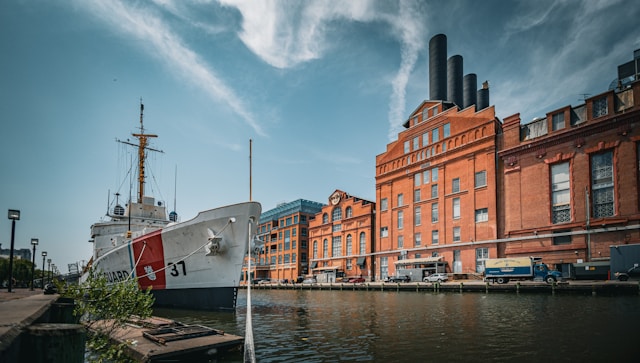 A waterfront view of Baltimore's Inner Harbor, featuring a Coast Guard ship, brick buildings, and industrial smokestacks against a blue sky with wispy clouds.