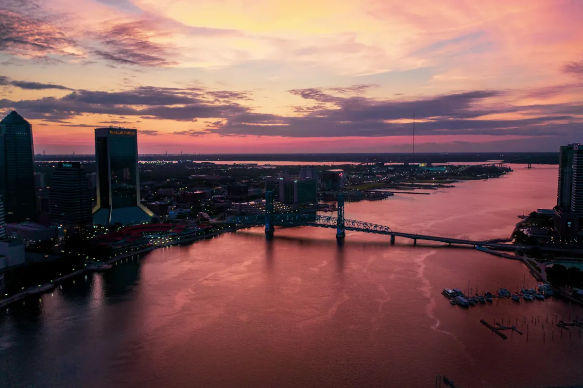 Jacksonville skyline at dusk with the Main Street Bridge illuminated in blue, reflecting off the St. Johns River, and the cityscape glowing under a vibrant sunset, intellectual property attorney services for small businesses.