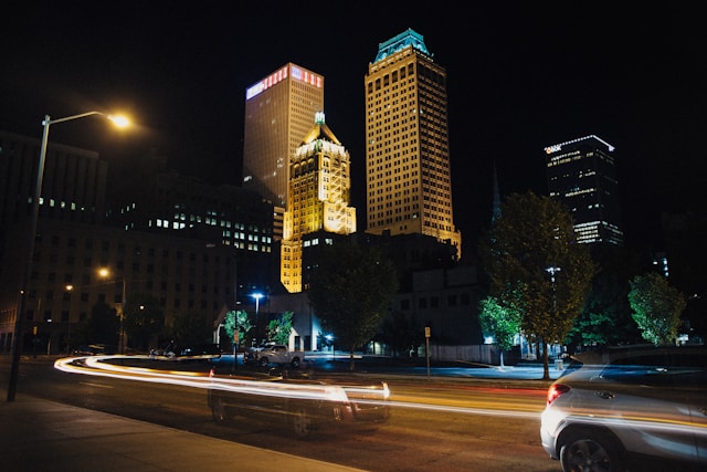 Nighttime skyline of downtown Tulsa, Oklahoma, featuring illuminated skyscrapers, light trails from traffic, and trees lining the street.