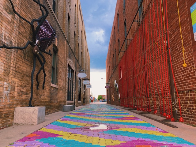 A colorful, rug-like design covering an alleyway in downtown Wichita, Kansas, with brick buildings on both sides and artistic sculptures.